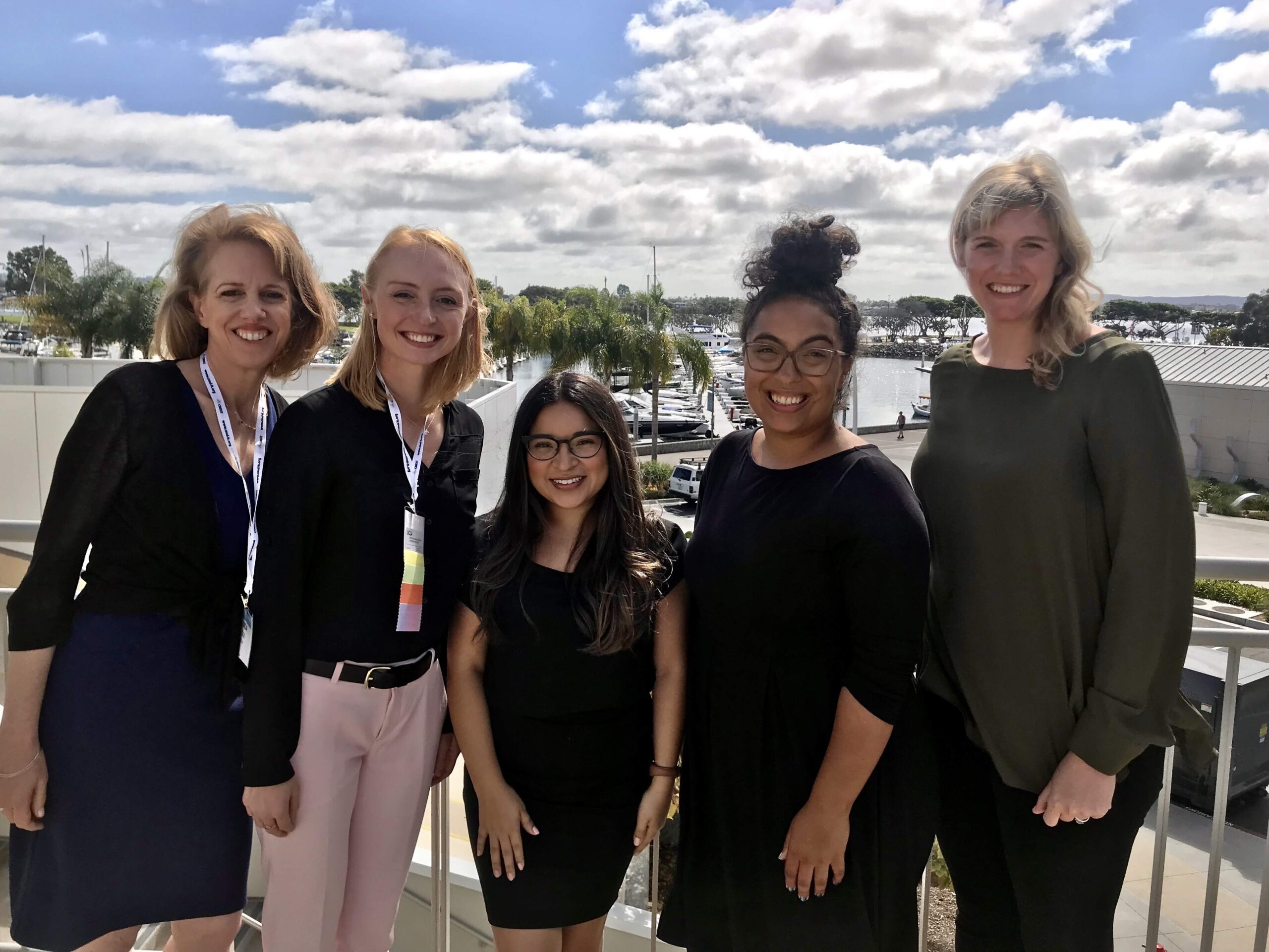 A photo of five members of the Momentum team in front of a marina. It is a mostly cloudy blue sky with palm trees in the background.