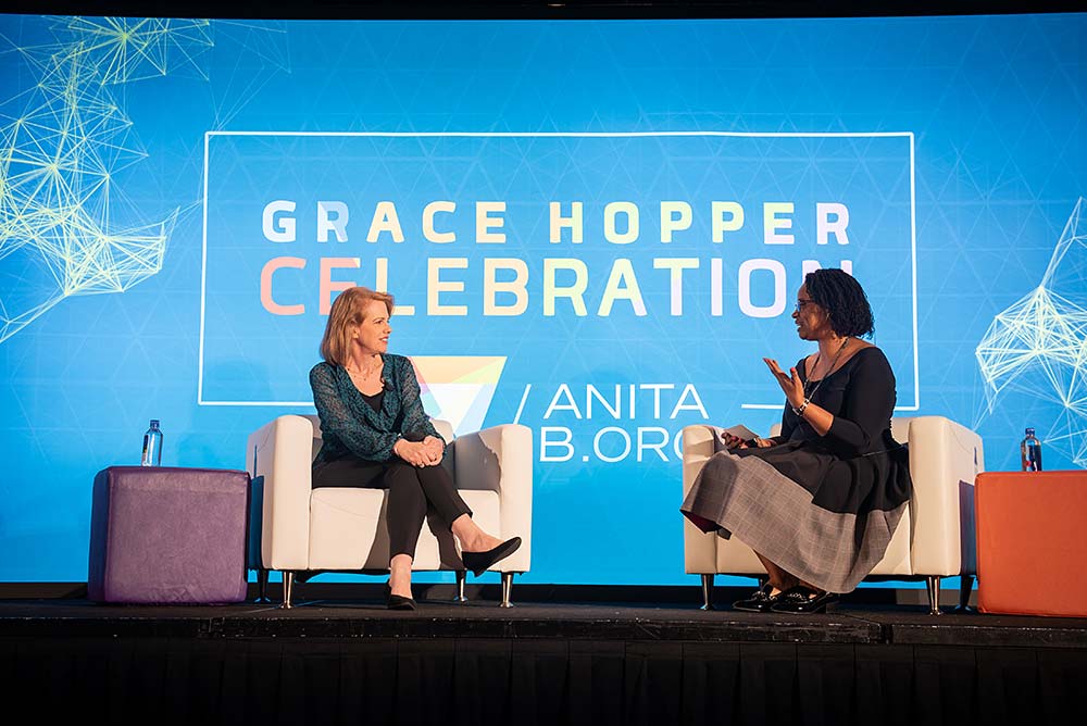 Dr. Linda Sax (left) discusses BRAID Research with AnitaB President and CEO, Brenda Darden Wilkerson (right) during the 2019 Grace Hopper Celebration. Linda and Brenda are seated in white cushioned seats, facing each other. In the background is a blue backdrop that reads: Grace Hopper Celebration.