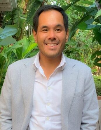 Man in white collared shirt and grey jacket in front of greenery.