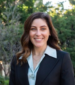 Katie Stormes, Ph.D., wearing a light blue blouse and dark blazer posing in front of greenery and trees in the sunlight.