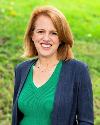 Close-up of a red-haired white woman, smiling, arms crossed, wearing a blue sweater with grass in the background.
